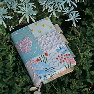 以下が含まれることがあります： 蝶の模様と花の刺繍が施された、青と白のパッチワークの小さなノート。ノートは緑の葉の上に置かれています。