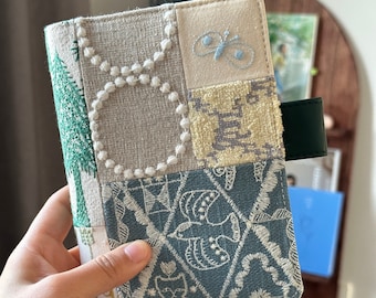 May include: A handmade fabric journal cover with a patchwork design. The cover features a variety of fabrics in shades of green, blue, and white, with embroidered details. The cover is secured with a green strap.