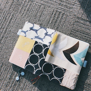 May include: A patchwork fabric notebook cover with a variety of patterns, including circles, geometric shapes, and animal prints. The cover is made of different colored fabrics, including black, white, blue, yellow, and pink.