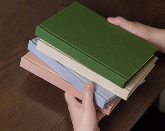 May include: A stack of four hardcover journals in forest green, cream, light blue, and dusty pink. The journals feature solid-colored covers and plain, off-white interior pages. One journal is shown open with a black pen resting on the page.