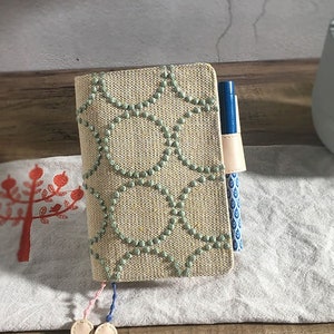 May include: A beige notebook cover with a green and white geometric pattern. The cover has a pen holder and a tassel.