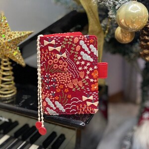 May include: A red fabric planner with a floral and cloud embroidery design. The planner has a white tassel and a red strap.
