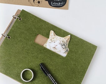 May include: A green felt-covered notebook with a cat sticker peeking out from a cut-out window. The notebook has a ring binder and a silver ring. A black pen and a silver ring are on the cover.