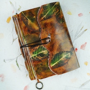 Può includere: Un diario in pelle marrone con un design a foglia verde e una chiusura a cinturino in pelle marrone. Il diario ha un aspetto rustico ed è perfetto per scrivere o tenere un diario.