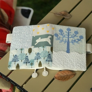 May include: A white fabric notebook cover with a patchwork design featuring blue trees, a white rabbit, and yellow stars. The cover has a white lace trim and a blue tassel.