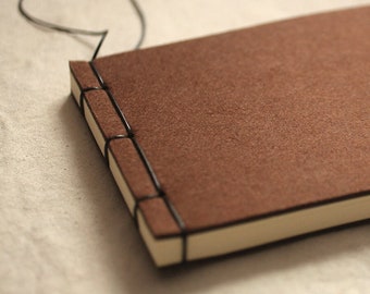 May include: A brown leather-bound journal with black stitching and a cream-colored page edge. The journal is closed and has a black string tied around it.