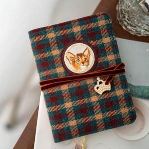 Puede incluir: Un diario a cuadros con un bordado de gato en la portada. Cuenta con un cierre de cinta de terciopelo marrón y un charm de corona decorativo. El patrón a cuadros incluye colores turquesa, marrón y beige.