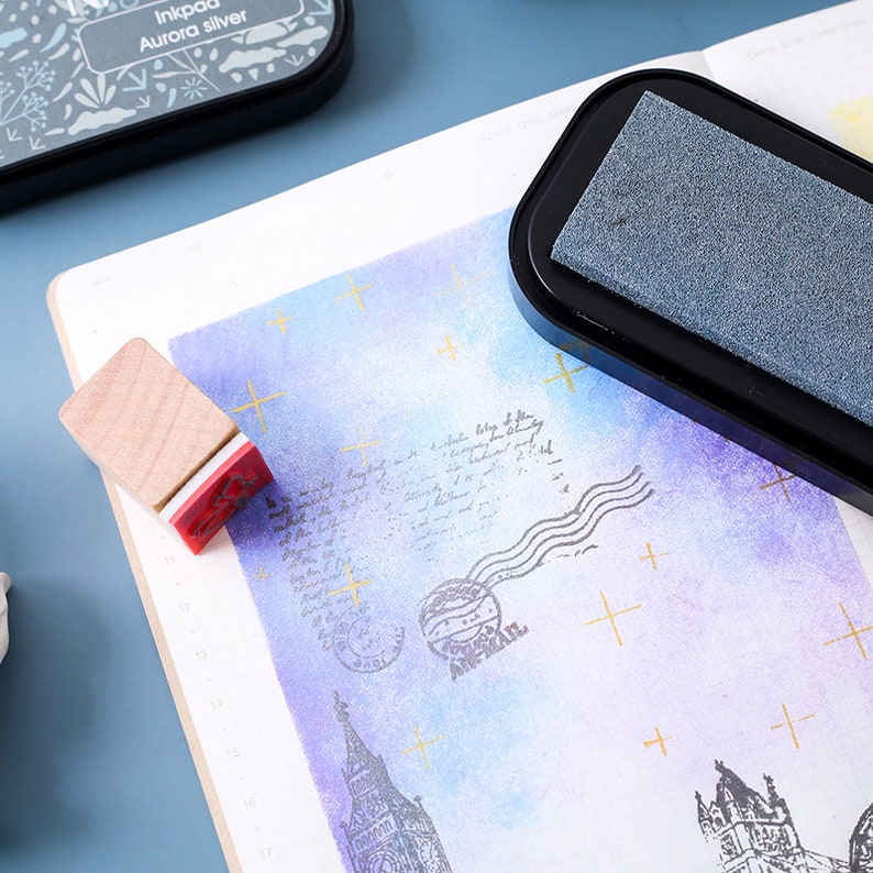 May include: A close-up of a black ink pad with a gray ink pad inside. The ink pad is on a white surface with a purple and blue watercolor background. There are gold crosses and a stamp of a building on the background.