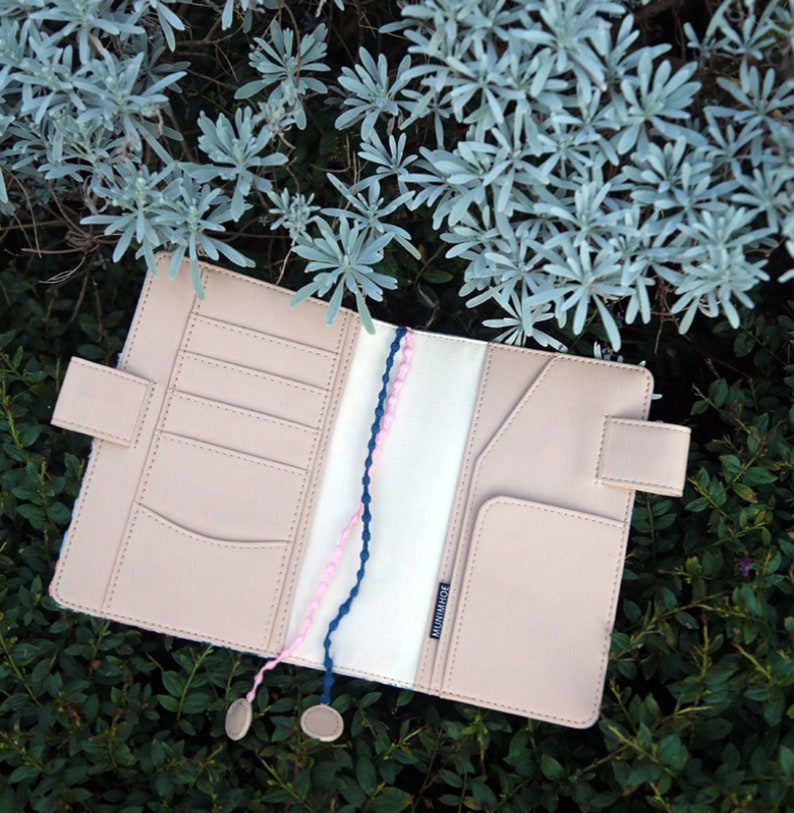 May include: An open beige leather planner with a white interior and two pink and blue braided cords. The planner has multiple card slots and a pocket on the right side. The planner has the word "MIDORI" printed on the interior.