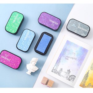 May include: Six different colored ink pads with the word "MAGNIFICOUS" printed on them. The ink pads are arranged on a light blue surface.