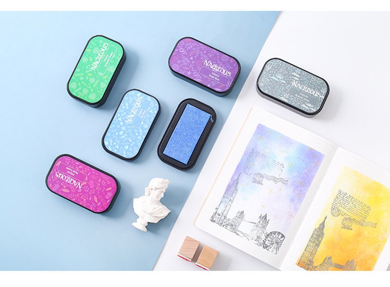 May include: Six different colored ink pads with the word "MAGNIFICOUS" printed on them. The ink pads are arranged on a light blue surface.