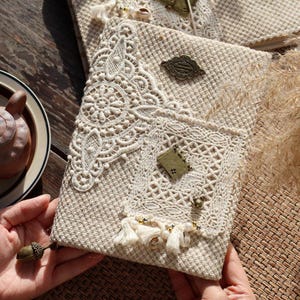 Peut inclure: Une couverture de journal en tissu beige ornée de superpositions de dentelle blanche complexe, d'une poche en dentelle rectangulaire et d'un emblème en métal bronze. Elle est décorée de petits pompons blancs, de perles et d'une breloque livre.