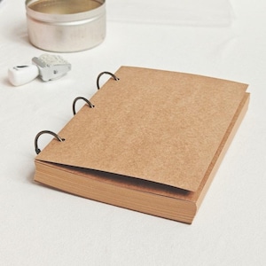 May include: A small, open kraft paper notebook with three metal rings. The notebook is on a white surface, with a small metal tin and a stamp nearby. The pages are a light brown colour.