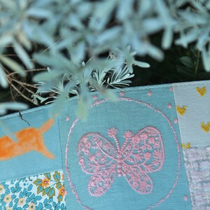 May include: A patchwork fabric with a pink embroidered butterfly in a circle on a light blue background. The fabric is made up of different patterns and colors, including orange, yellow, and white.