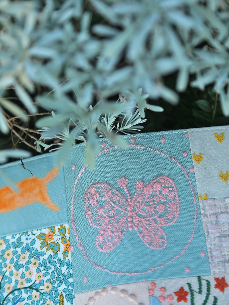 May include: A patchwork fabric with a pink embroidered butterfly in a circle on a light blue background. The fabric is made up of different patterns and colors, including orange, yellow, and white.