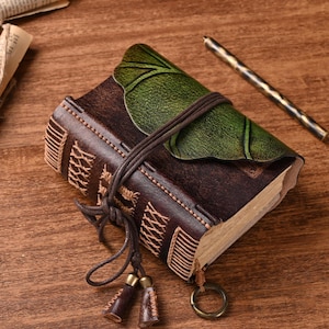 May include: A handmade leather journal with a green leaf-shaped cover and a brown leather strap. The journal features decorative stitching and a tassel with two small brown tassels. A pen rests on the wooden surface.