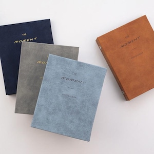 May include: Four photo albums in different colours: navy blue, grey, light blue, and brown. Each album has the text "The Moment" and "Photo Album" printed on the front.