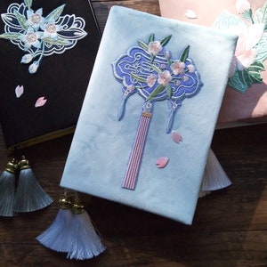 May include: Three embroidered journals with floral designs. The first journal is black with a white floral design. The second journal is light blue with a pink and white floral design. The third journal is pink with a green and white floral design.