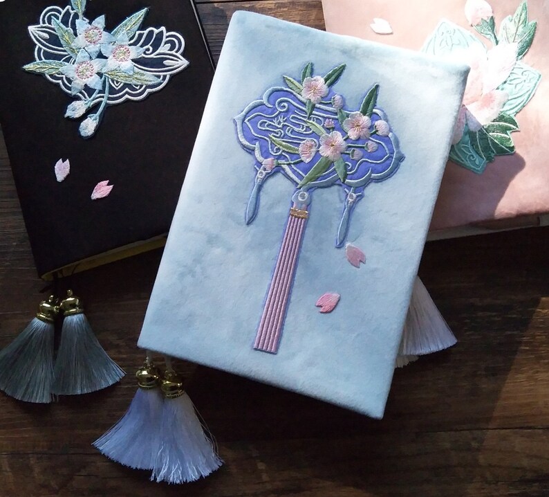 May include: Three embroidered journals with floral designs. The first journal is black with a white floral design. The second journal is light blue with a pink and white floral design. The third journal is pink with a green and white floral design.