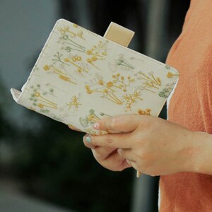 May include: A white fabric notebook with a floral embroidery design in yellow and green. The notebook has a tan leather closure tab.