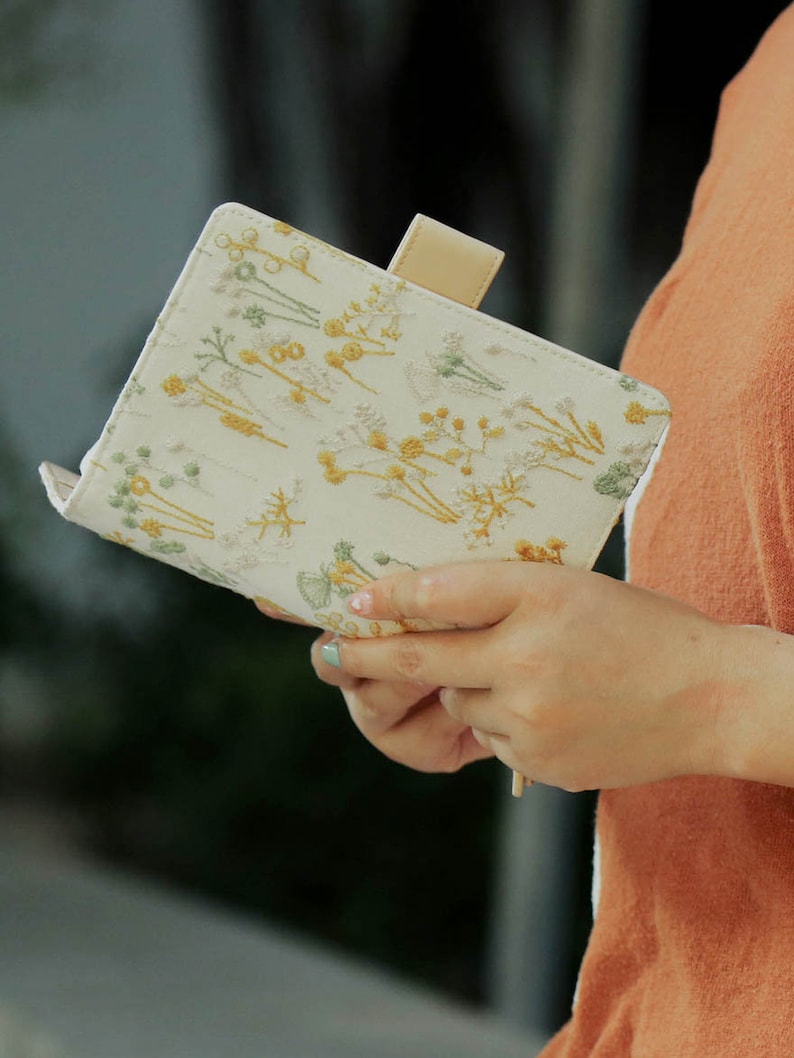 May include: A white fabric notebook with a floral embroidery design in yellow and green. The notebook has a tan leather closure tab.