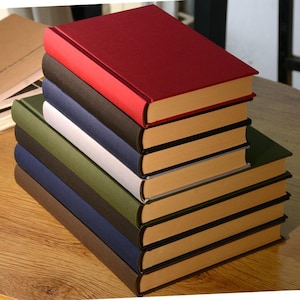 Puede incluir: Una pila de ocho libros encuadernados con cubiertas de diferentes colores. El libro de arriba es rojo, el segundo es negro, el tercero es azul marino, el cuarto es blanco, el quinto es verde, el sexto es azul oscuro, el s&eacute;ptimo es marr&oacute;n y el octavo es beige.