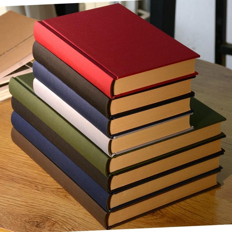 Puede incluir: Una pila de ocho libros encuadernados con cubiertas de diferentes colores. El libro de arriba es rojo, el segundo es negro, el tercero es azul marino, el cuarto es blanco, el quinto es verde, el sexto es azul oscuro, el s&eacute;ptimo es marr&oacute;n y el octavo es beige.