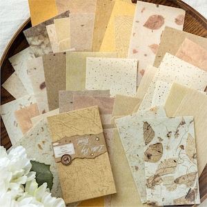 May include: A selection of handmade paper in beige, brown, and yellow hues, featuring diverse textures and leaf details. A small booklet with the text "Letter By Word" is also present. The papers are arranged in a wooden bowl.