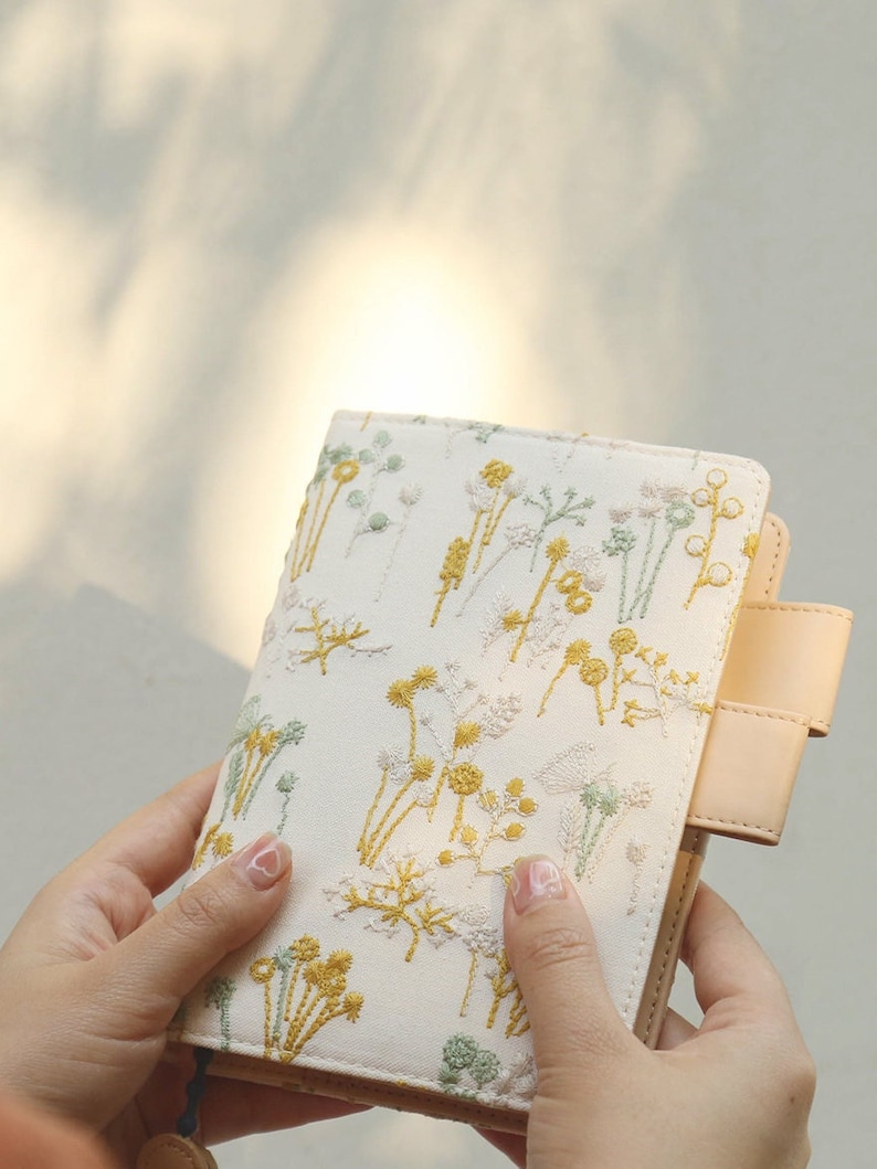 May include: A white fabric notebook cover with a floral embroidery design in yellow and green. The cover has a beige leather flap with a strap.