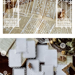 May include: A set of eight white paper cards with a delicate lace border. The cards are arranged on a brown surface with a book and other decorative items.