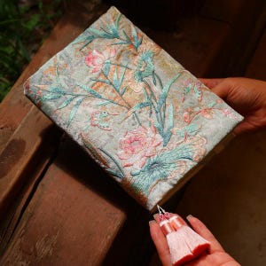 May include: A rectangular journal with a light blue velvet cover featuring intricate embroidery of pink lotus flowers, green leaves, and stems. The book includes a decorative light pink double tassel attached to the bottom edge with a small white cord.