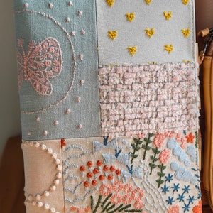 May include: A light blue, beige, and pink fabric notebook cover with embroidered flowers, hearts, and stars. The cover is made of patchwork fabric and has a unique, handmade look.