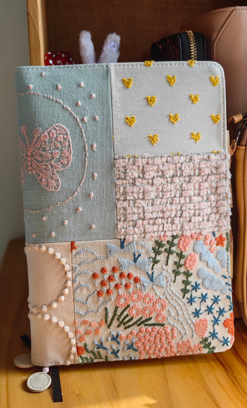 May include: A light blue, beige, and pink fabric notebook cover with embroidered flowers, hearts, and stars. The cover is made of patchwork fabric and has a unique, handmade look.
