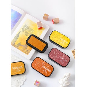 May include: Four ink pads with different colored inks and patterns. The ink pads are labeled "Nacreous" and have the ink color listed below. The ink colors are Galaxy Gold, Summer Rust, Cosmic Copper, and Autumn Leaves.