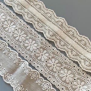 May include: Three different styles of white cotton lace trim with a delicate floral pattern. The lace is on a beige background.
