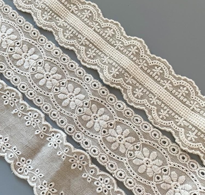 May include: Three different styles of white cotton lace trim with a delicate floral pattern. The lace is on a beige background.