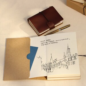 May include: A brown leather bound journal with a brown paper envelope attached to the front cover. The journal is open to a page with a black and white ink drawing of a building. The journal has a black string closure.