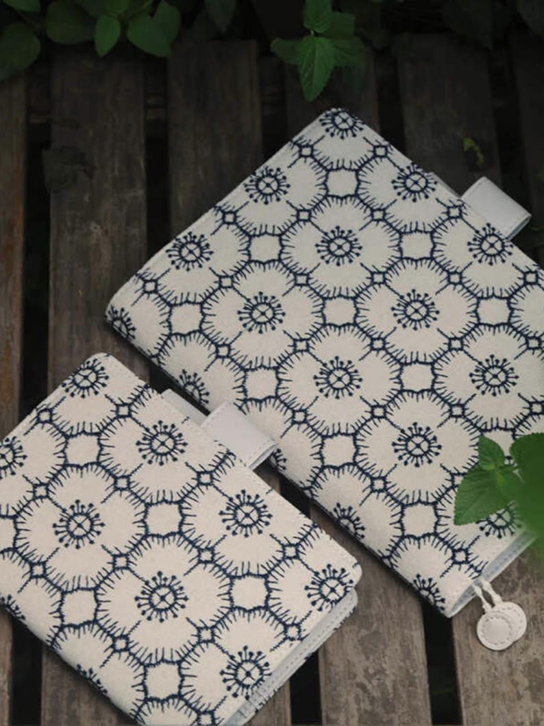 Op de afbeelding: Twee witte stoffen notitieboekhoezen met een blauw geometrisch patroon. De hoezen hebben een witte flap met een witte knop.