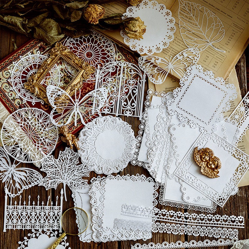 May include: Assortment of white lace paper doilies with various designs, including butterflies, gates, and leaves. The doilies are arranged on a wooden surface with a book and other decorative items.