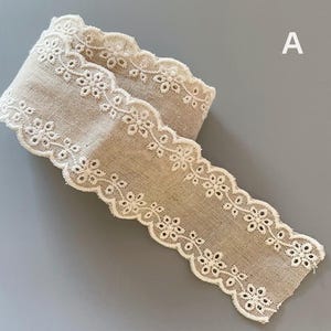 May include: A beige cotton lace trim with a scalloped edge and white embroidery. The trim is folded in half and the edges are visible.