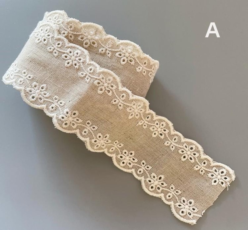 May include: A beige cotton lace trim with a scalloped edge and white embroidery. The trim is folded in half and the edges are visible.