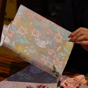 May include: A rectangular fabric pouch featuring a shimmering, iridescent finish with a woven pattern of light pink lilies, teal leaves, and small white cat figures. The surface is accented with tiny gold star motifs throughout the floral design.