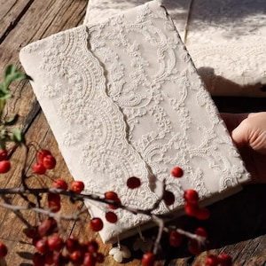May include: Ivory-colored journal with intricate lace overlay. The journal's cover features a detailed floral lace pattern, with a scalloped edge. The journal is held near a branch with red berries, suggesting a natural theme.