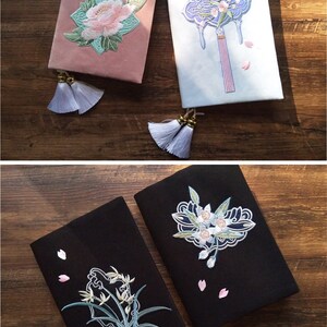 May include: Six different book covers with embroidered designs. The covers are in various colors, including black, white, blue, pink, and green. Each cover features a different floral or nature-inspired design, such as a crane, bamboo, a flower, and a fan. The covers have tassels attached to them.