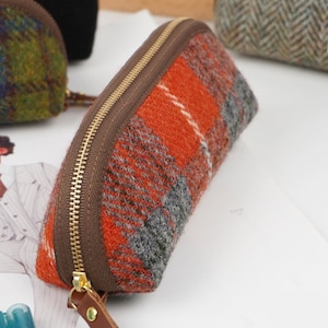 May include: A semi-circular pencil case made of orange, gray, and white plaid wool fabric. The case features a brown fabric trim along the top edge, a gold-toned metal zipper, and a brown leather pull tab attached to the zipper slider.