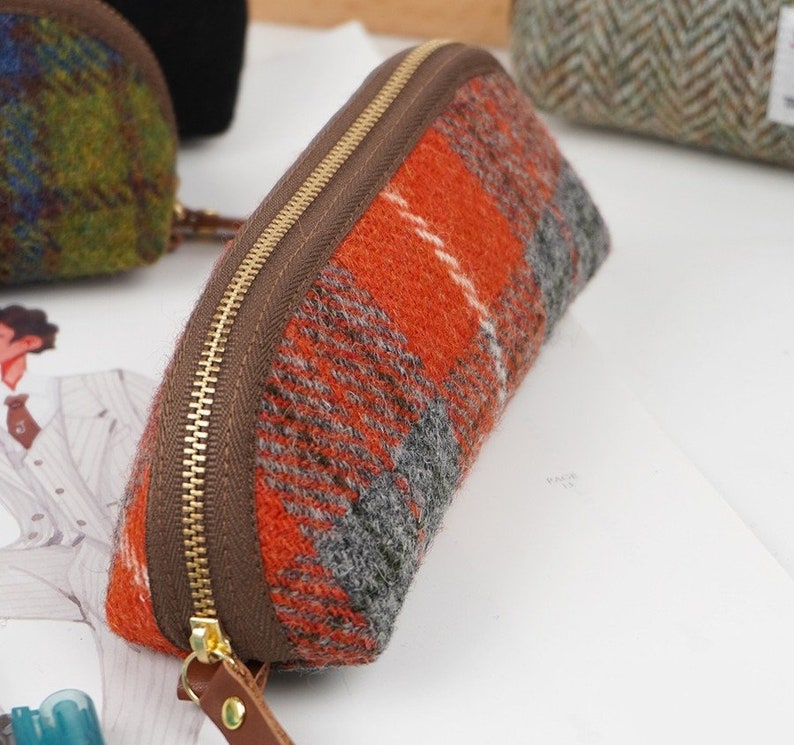 May include: A semi-circular pencil case made of orange, gray, and white plaid wool fabric. The case features a brown fabric trim along the top edge, a gold-toned metal zipper, and a brown leather pull tab attached to the zipper slider.