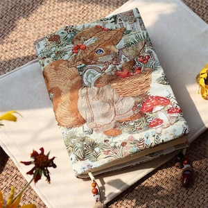 May include: A rectangular notebook with a woven fabric cover featuring a squirrel wearing a hat and carrying a basket of red berries. The scene includes red and white mushrooms, green foliage, and a small house. A beaded bookmark hangs from the bottom.