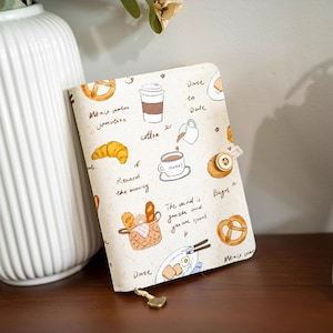 Può includere: Una copertina per quaderno di colore crema con un design fantasioso che presenta tazze di caffè, pasticcini e testo scritto a mano. Il testo include frasi come "caffè", "registrare la bellezza" e "il vento è gentile".