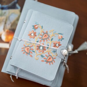May include: A stack of pale blue notebooks featuring floral embroidery in orange, teal, and gold. The smallest notebook has a pearl button closure and a tassel. A white string wraps around the notebooks.