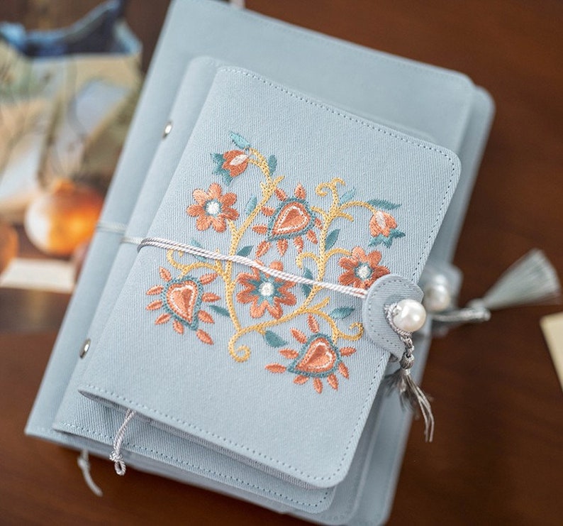 May include: A stack of pale blue notebooks featuring floral embroidery in orange, teal, and gold. The smallest notebook has a pearl button closure and a tassel. A white string wraps around the notebooks.